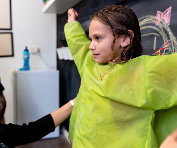 Young girl getting help putting on her art smock at the Voice Within Therapy Centre